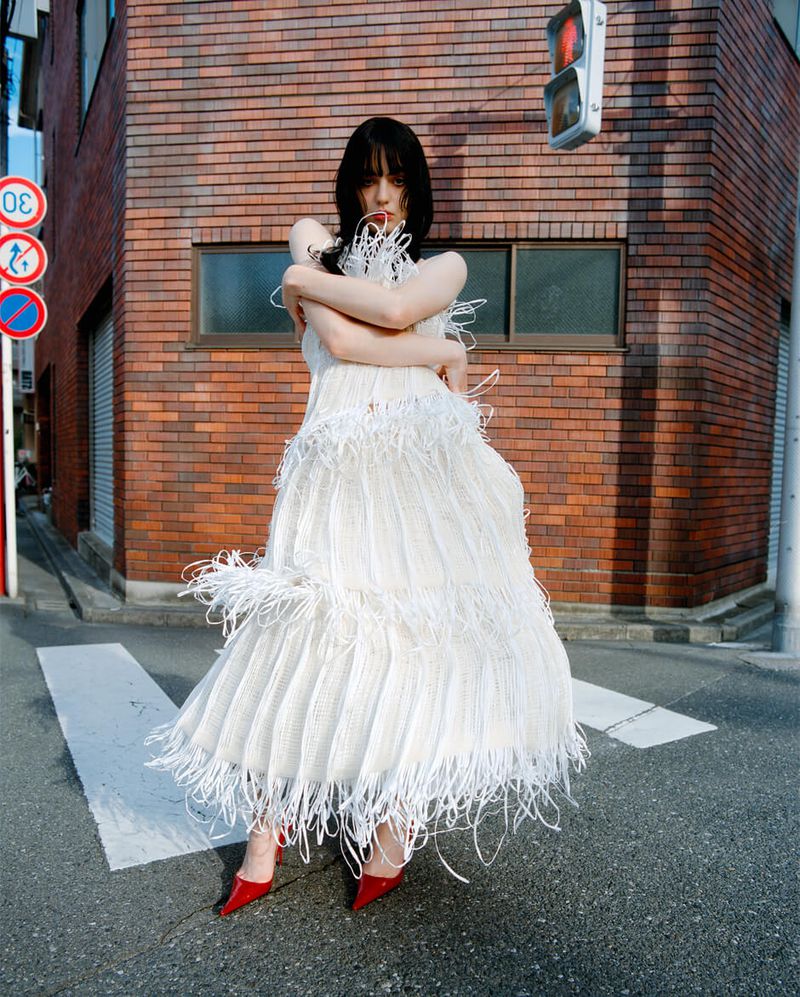 A young adult female stands on a street in a white feather dress and red high heels, with a brick building in the background.
