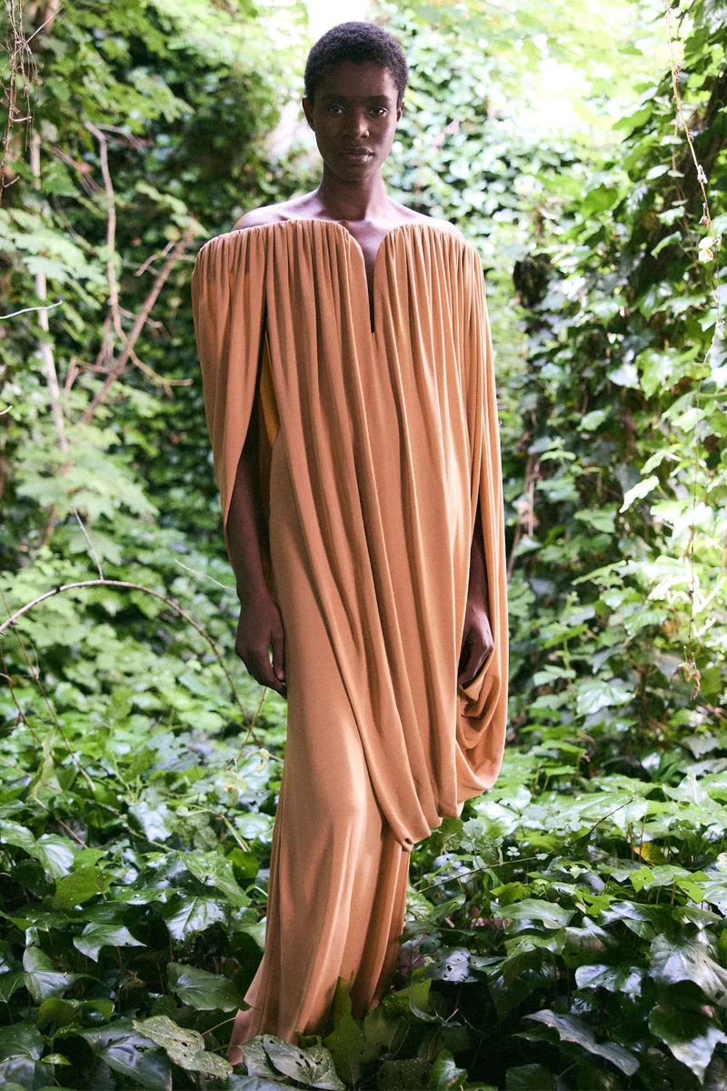 An adult fashion model in a flowing light-brown gown stands amidst a lush green forest of ivy and foliage in an outdoor setting.