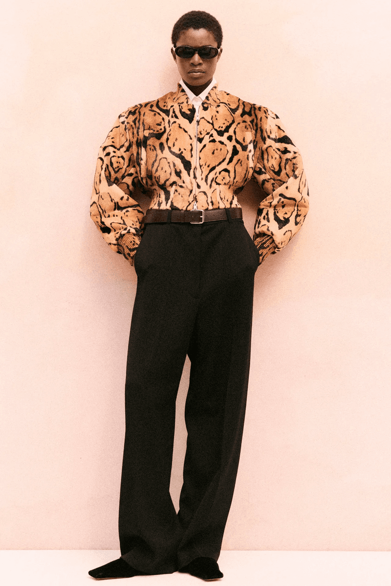 A fashion model poses in a studio, wearing a printed blouse with shoulder details, high-waisted black pants, black sunglasses and black flats.