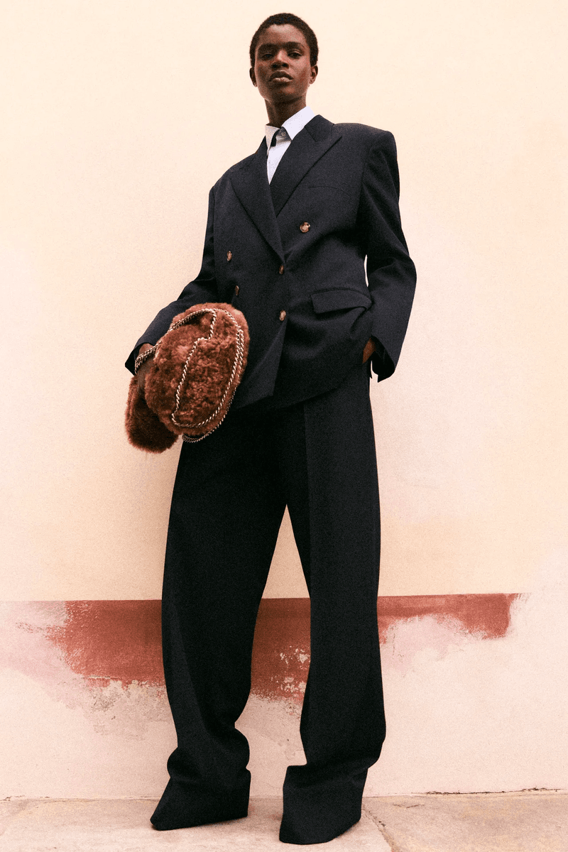 An African-American woman in oversized black suit, with a white shirt and tie, holding a brown fur bag, standing against peach wall.