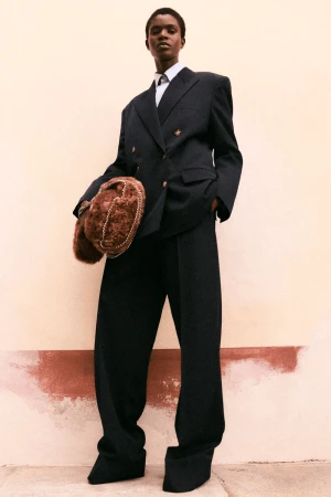 An African-American woman in oversized black suit, with a white shirt and tie, holding a brown fur bag, standing against peach wall.