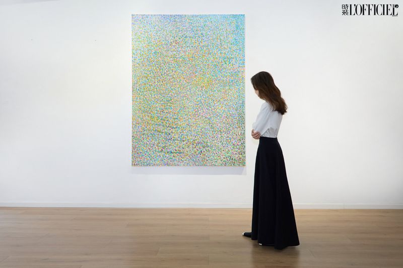 Woman views pointillist painting with blue, green, and yellow hues in a white-walled art gallery setting with wood flooring.