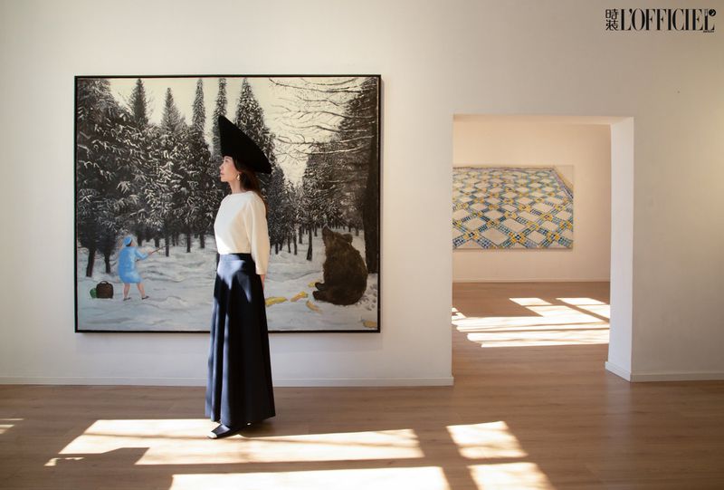Woman in hat standing in art gallery with winter landscape painting featuring a child, bear, and snow-covered trees.