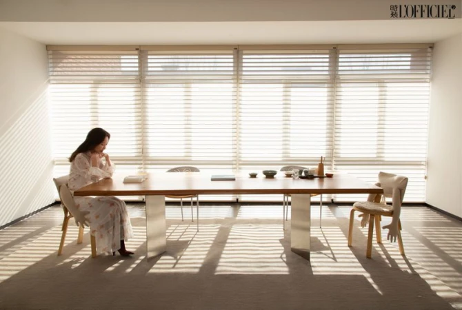 Woman in a floral dress sits at a long wooden table with decor and chairs in a naturally lit white room with sunlight shadows.