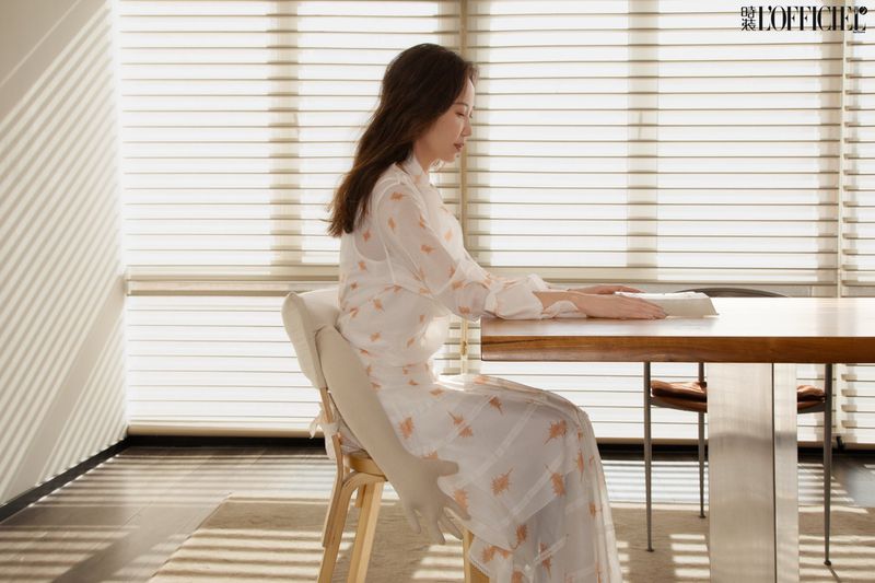 A woman in a floral dress sits at a light wood table in front of a window with blinds. The room is brightly lit with natural light.