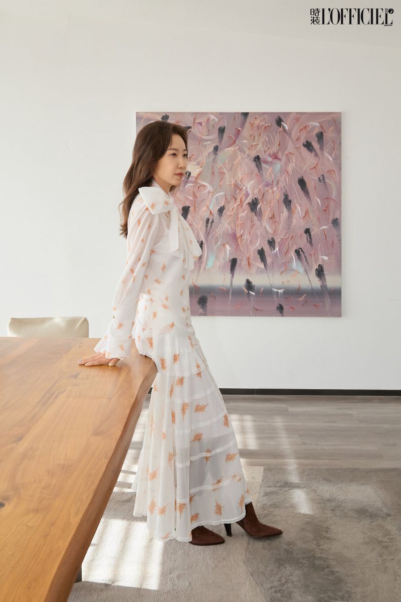 Woman in a long white dress poses next to a wooden table in an indoor setting with a painting on the wall. Fashion editorial.