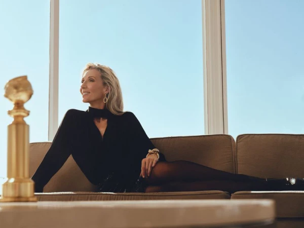 Elegant woman lounges on a couch in front of a large window, smiling, with the Golden Globe statue in the foreground. Indoor, daytime.