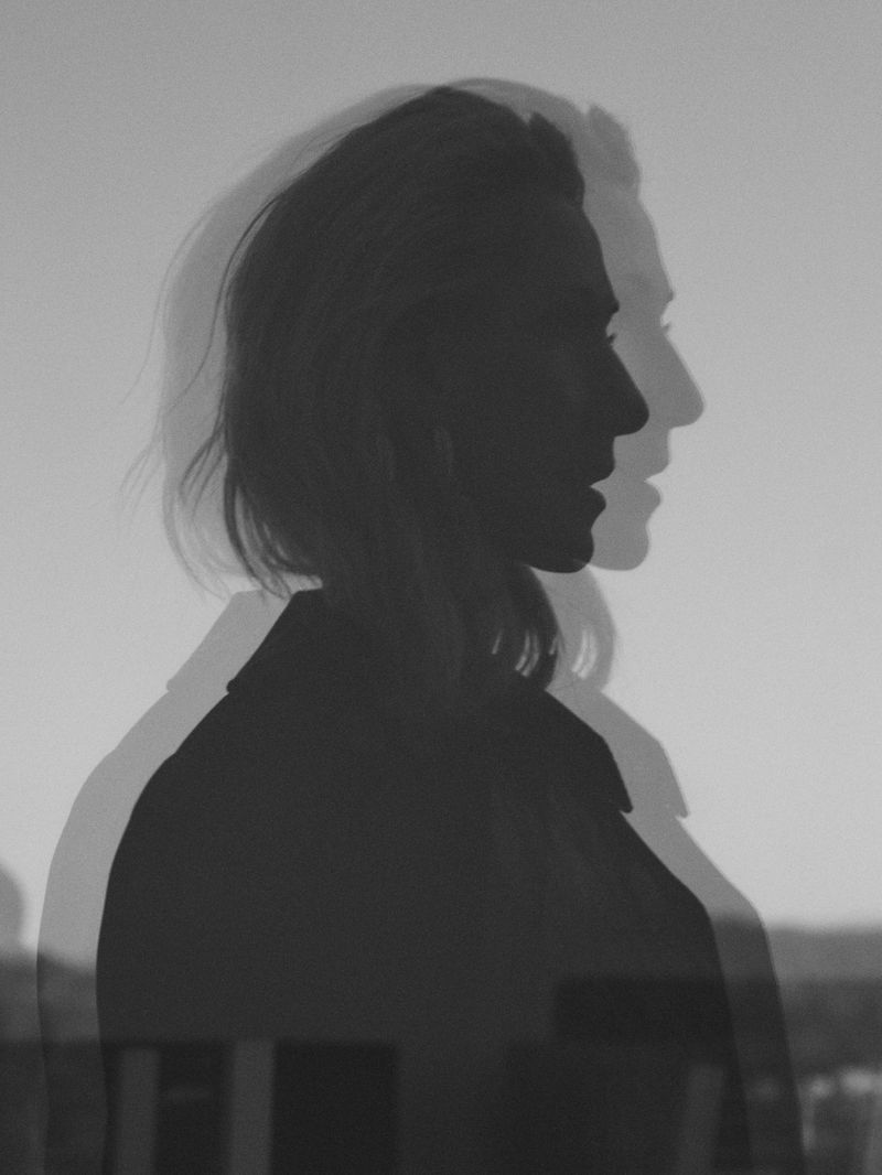 Monochromatic portrait of an adult female with a double exposure effect, featuring the side profile silhouette of her head and shoulder.