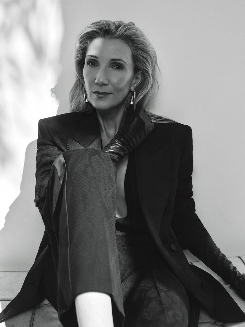 Helen Hoehne in a sharp black tailored blazer, dark leather gloves, and silver hoop earrings against a neutral, light-dappled wall.