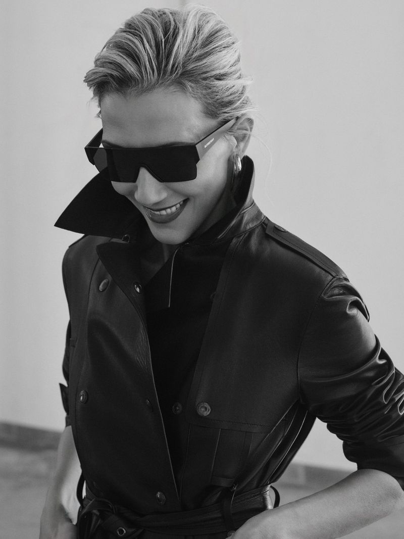 Portrait of adult woman in black leather trench coat and oversized black sunglasses against a neutral studio backdrop; fashionable and modern.