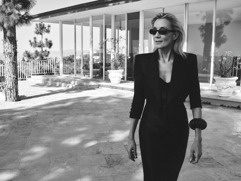 Elegant woman in black dress and sunglasses stands on a stone patio in front of a modern house with glass walls and a tree, black and white.