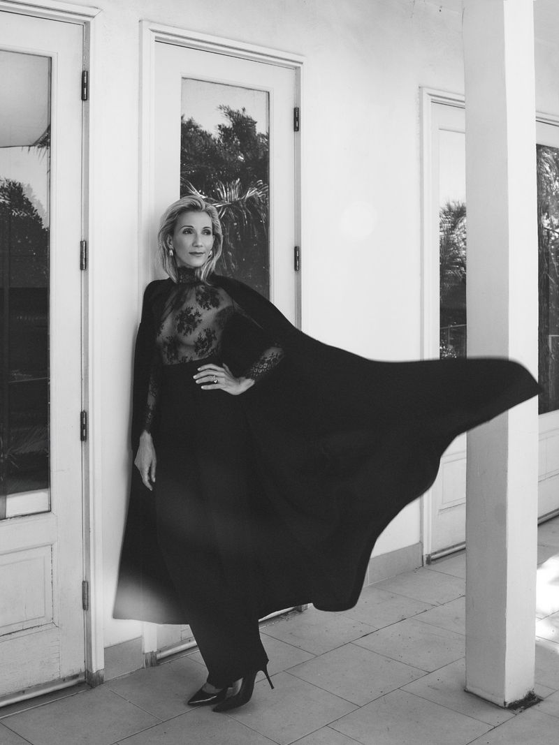 Elegant woman in a black lace dress with a flowing cape poses against a white building, showcasing a blend of classic style and modern fashion.