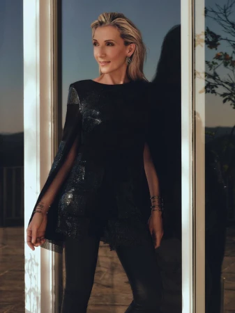 Elegant blonde woman in a sequined top poses confidently in front of a modern window, showcasing fashion and style in a lifestyle setting.
