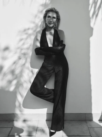 Monochrome portrait of a woman in a black halter jumpsuit and gloves leaning against a light wall with palm shadows.