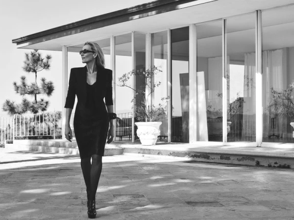 Woman wearing a dark fitted dress and blazer walking outdoors in front of a modern house exterior with large glass windows.