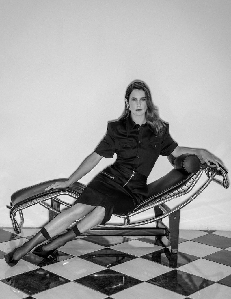 Woman in a short-sleeved black dress and sheer socks reclines on a metal chaise lounge against a white backdrop and diamond-patterned floor.