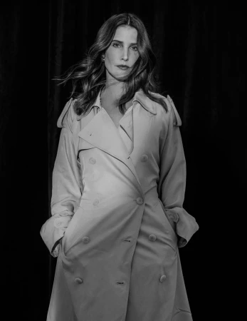 Monochrome portrait of actress Cobie Smulders wearing a double-breasted trench coat against a dark curtain background.