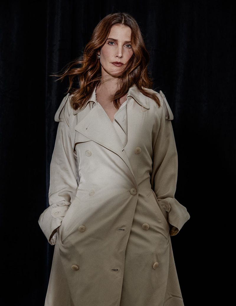 Cobie Smulders wearing a beige trench coat against a dark curtain background for Numero Netherlands.