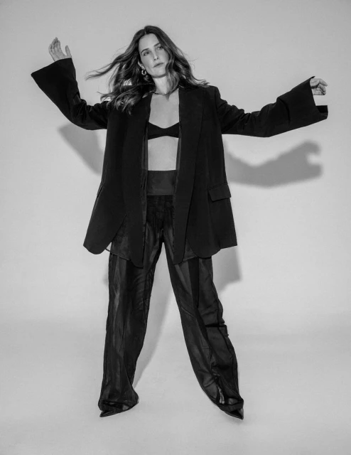 A full-length studio shot in black and white features a young woman in a bra, blazer, and trousers striking a dynamic pose against a white backdrop.