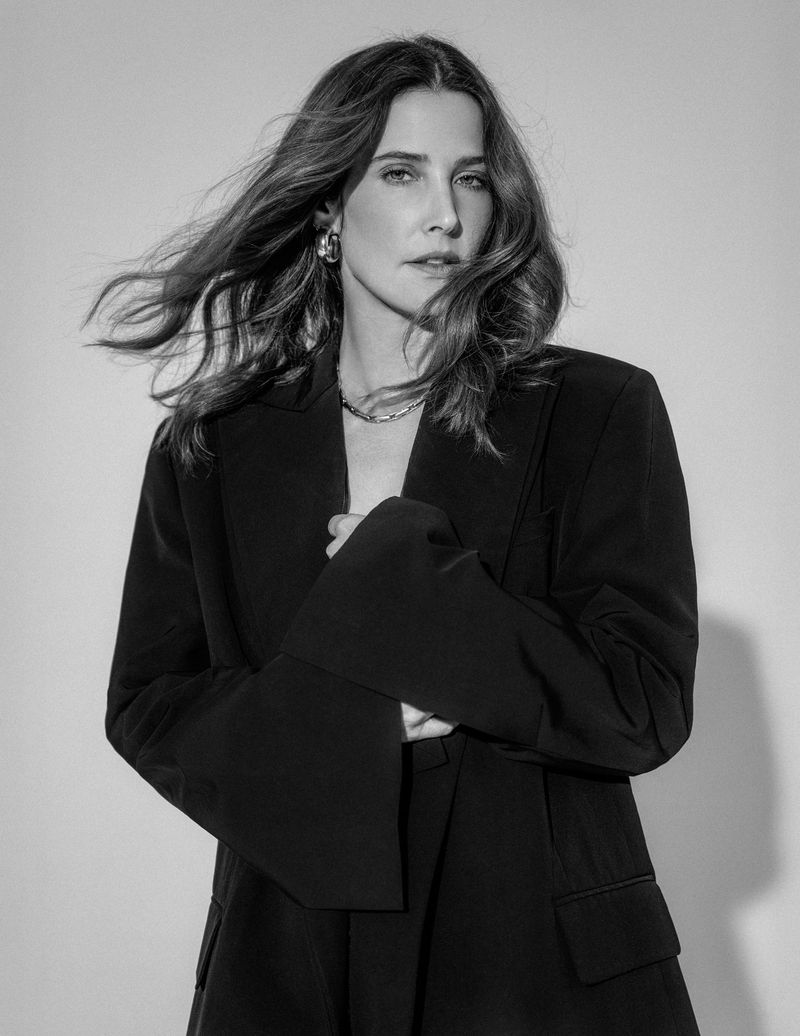 Black and white portrait of adult female posing in a dark blazer with loose hair, wearing jewelry and a neutral facial expression.