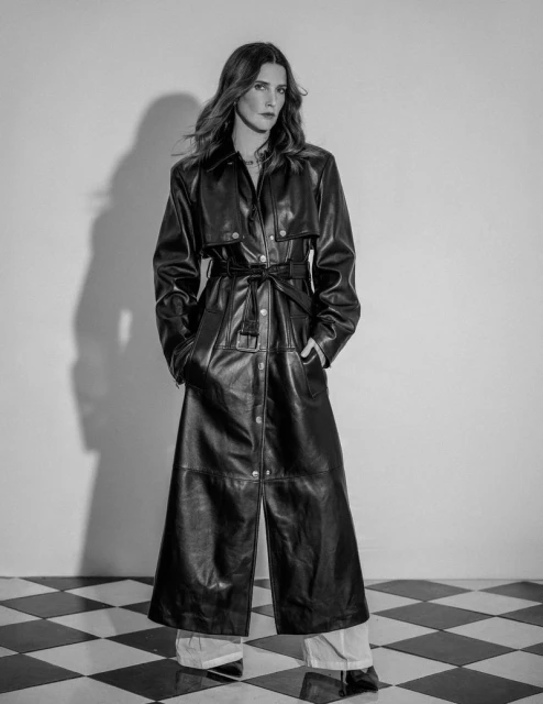 A woman with long brunette hair stands in a long black leather coat and light pants in a black and white studio shot.