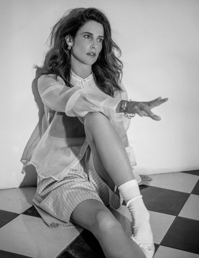 Black and white portrait of a woman sitting on a checkered floor wearing a sheer top, striped shorts, jewelry, and white socks.