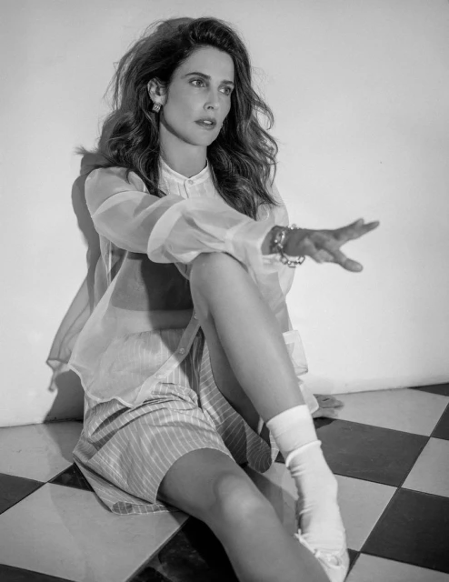 Black and white portrait of a woman sitting on a checkered floor wearing a sheer top, striped shorts, jewelry, and white socks.