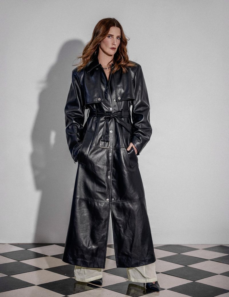 An adult woman poses in a full-length black leather trench coat with light pants and black shoes against a black-and-white checkered floor.