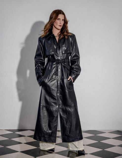 An adult woman poses in a full-length black leather trench coat with light pants and black shoes against a black-and-white checkered floor.