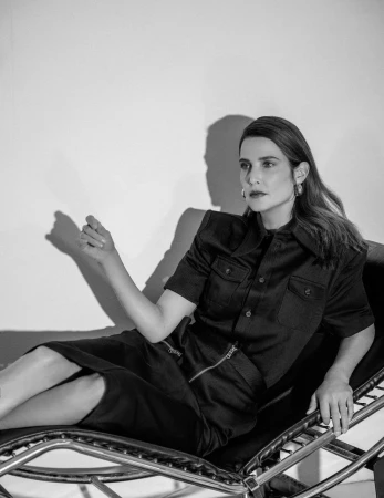 Cobie Smulders wearing a structured black short-sleeved collared shirt and pencil skirt, reclining on a lounge chair against a white wall.