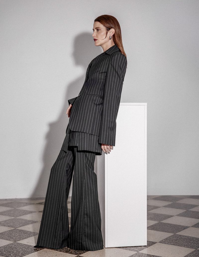 Cobie Smulders leaning against a white pedestal wearing a pinstripe pantsuit with wide-leg trousers