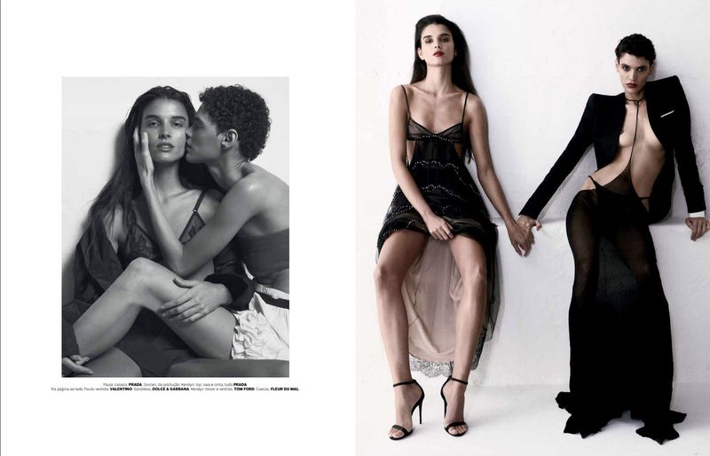 Two women in elegant black dresses hold hands against a white backdrop in this fashion editorial shot; one woman is being kissed on the cheek.