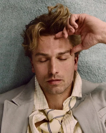 Close-up portrait of a man with his eyes closed, wearing a grid-patterned collared shirt and jacket against a textured blue wall.
