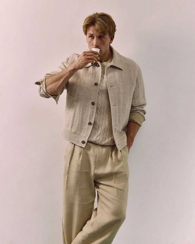 Blond male model in beige linen suit stands against neutral background, taking a sip from a small white paper cup, lifestyle fashion editorial.