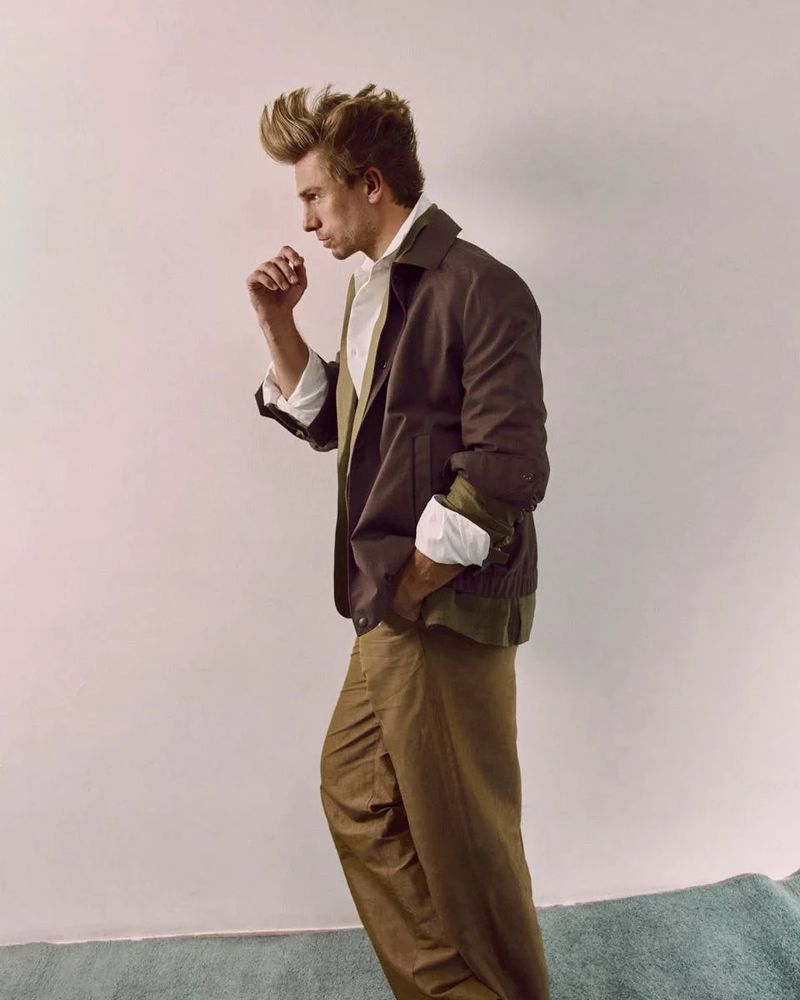 Studio shot of a male model with a distinctive hairstyle posing in a brown jacket and tan pants against a plain wall backdrop.