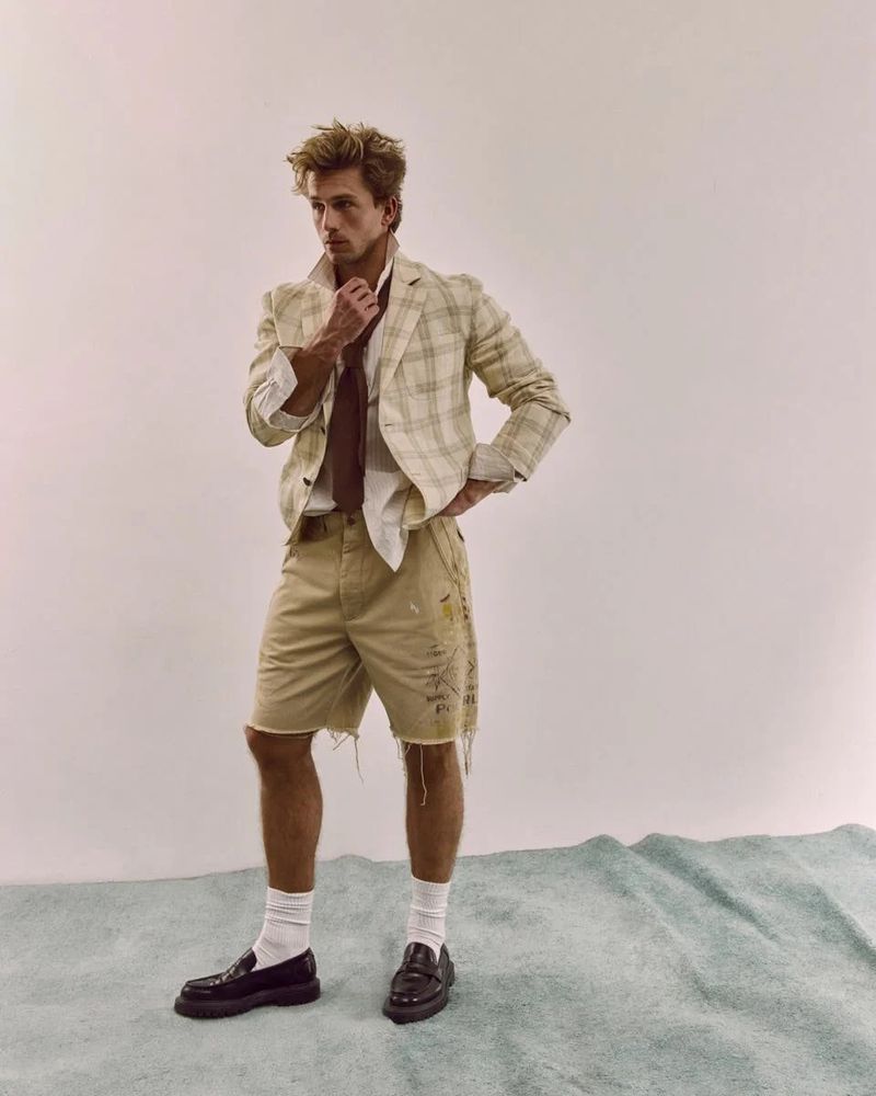 A stylish adult male poses in a studio wearing a plaid jacket, cutoff shorts, white socks, and loafers against a light backdrop.