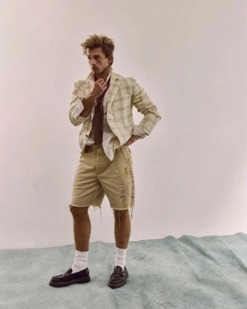 A stylish adult male poses in a studio wearing a plaid jacket, cutoff shorts, white socks, and loafers against a light backdrop.