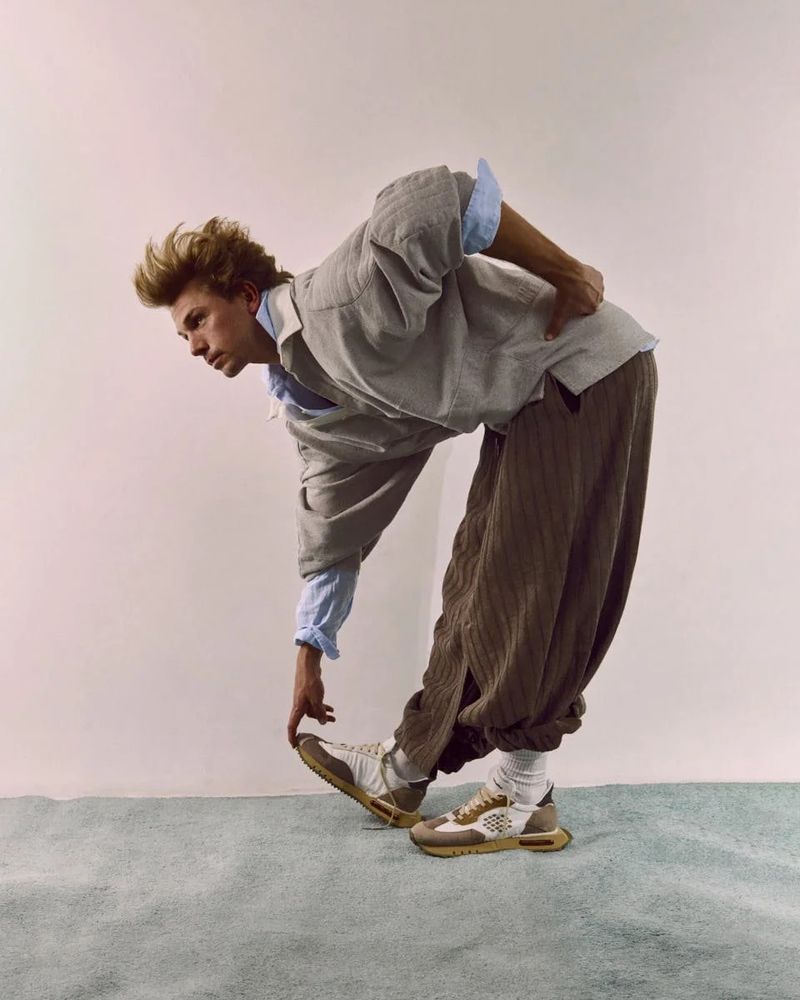 An adult male stretches in a photo studio wearing a light grey linen jacket, light blue button-down shirt, brown harem pants, and sneakers.