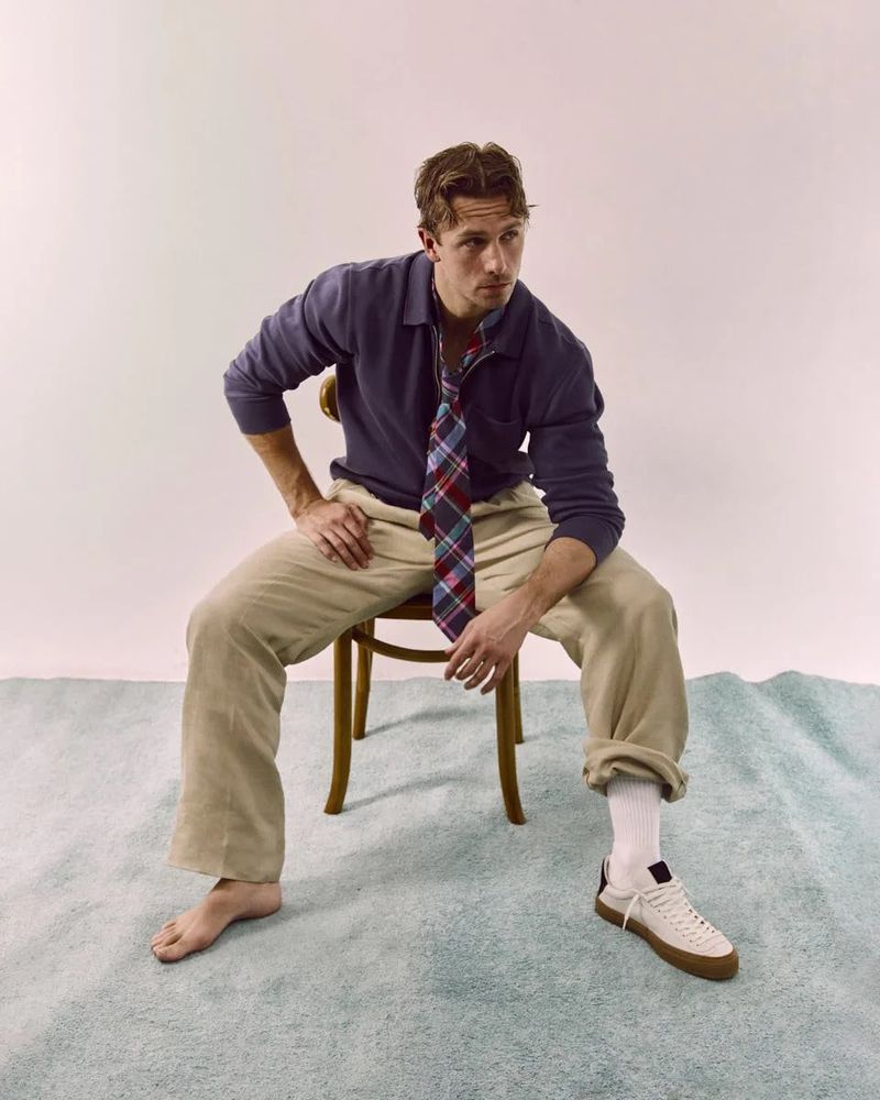 Man sits on wooden chair in relaxed pose with tie undone. He wears a blue shirt, cuffed pants, white socks, and sneakers on a textured surface.