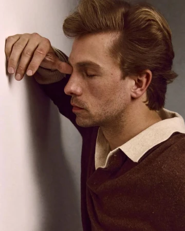 Profile of a man with eyes closed, leaning against a white wall, wearing a brown sweater with a cream-colored collar in soft lighting.