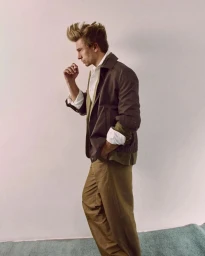 Profile portrait of a male model with styled hair wearing a dark brown jacket and khaki trousers against a plain wall.