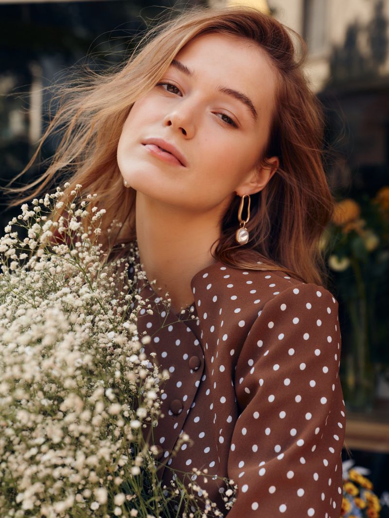 Close-up portrait of a woman with blonde hair holding white flowers wearing a brown polka dot dress.