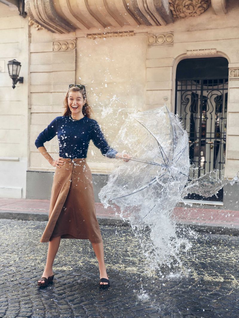 Happy woman in skirt and sweater stands on cobblestones as water explodes from an umbrella held in her right hand outside on a sunny day.