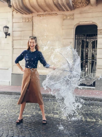 Happy woman in skirt and sweater stands on cobblestones as water explodes from an umbrella held in her right hand outside on a sunny day.