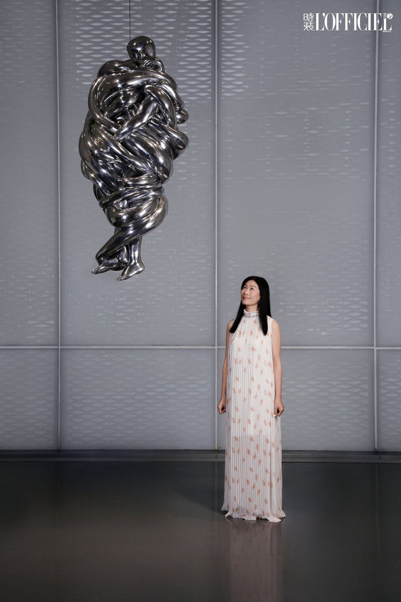 A long shot captures a woman in a patterned white dress standing in front of a metal art sculpture in an indoor gallery setting.