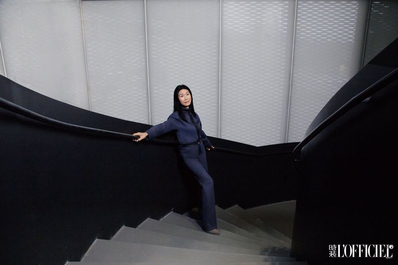 Adult woman in a blue pantsuit poses on grey stairs with black railings, set against a modern white wall with textured panels.