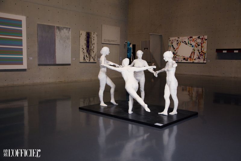 Art gallery with a sculpture of five white figures holding hands in a circle, surrounded by colorful abstract paintings on display.