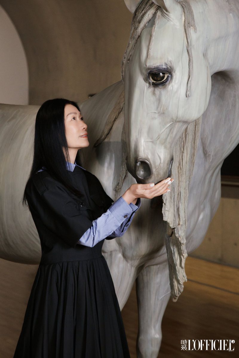 Adult female poses with a light grey horse statue indoors, wearing a black dress over a blue shirt; shot for L'Officiel magazine.
