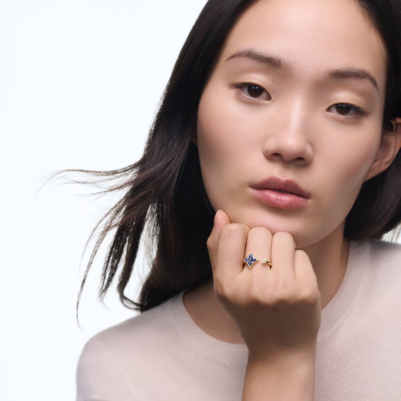 An Asian woman with dark hair poses indoors, with a delicate floral ring with a sapphire gemstone as the primary subject.