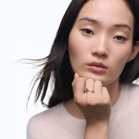 An Asian woman with dark hair poses indoors, with a delicate floral ring with a sapphire gemstone as the primary subject.